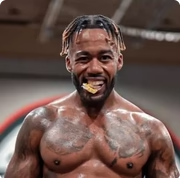 A close-up of a muscular, tattooed man with braided hair smiling with something on his tongue.