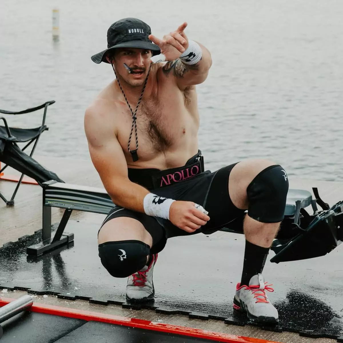 A shirtless man in a bucket hat and athletic gear crouches on a wet dock and points forward.