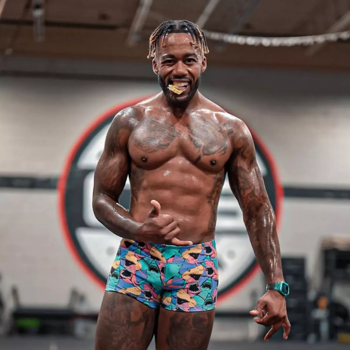 A muscular, tattooed man in colorful shark-print shorts smiles and makes a hand gesture in a gym.