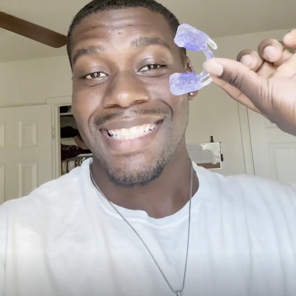 A smiling man in a white t-shirt holds up a clear, purple-tinted mouthguard.