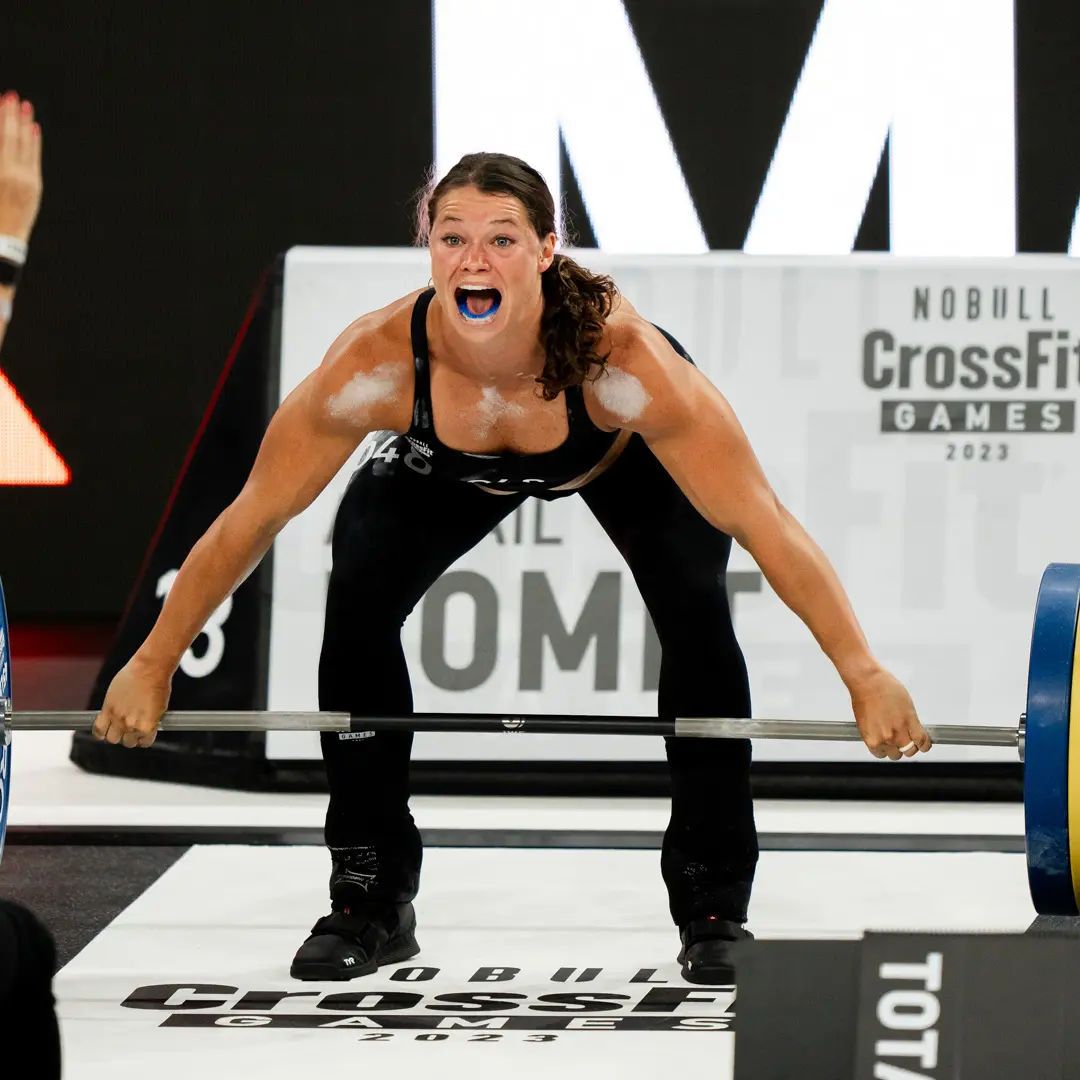 A female weightlifter with a blue mouthguard yells as she grips a heavy barbell during a competition.