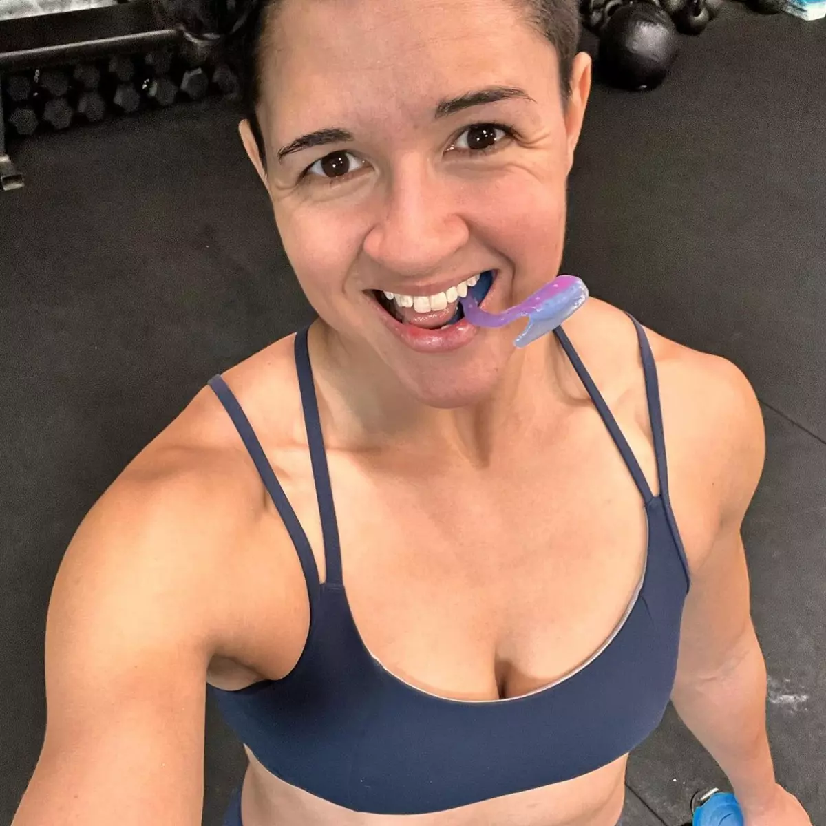 A smiling woman in a blue sports bra at a gym holds a purple whale-shaped object in her mouth.