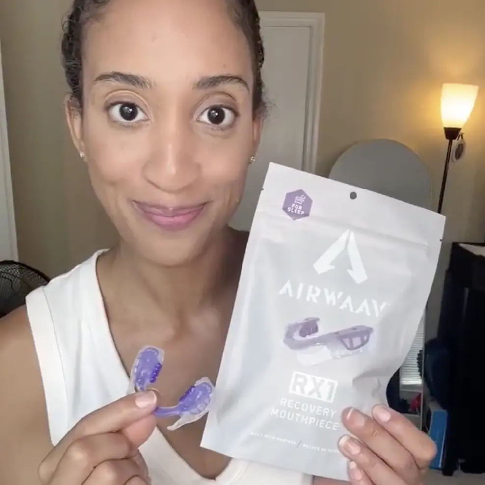 A woman holds up a purple recovery mouthpiece and its white packaging while smiling at the camera.