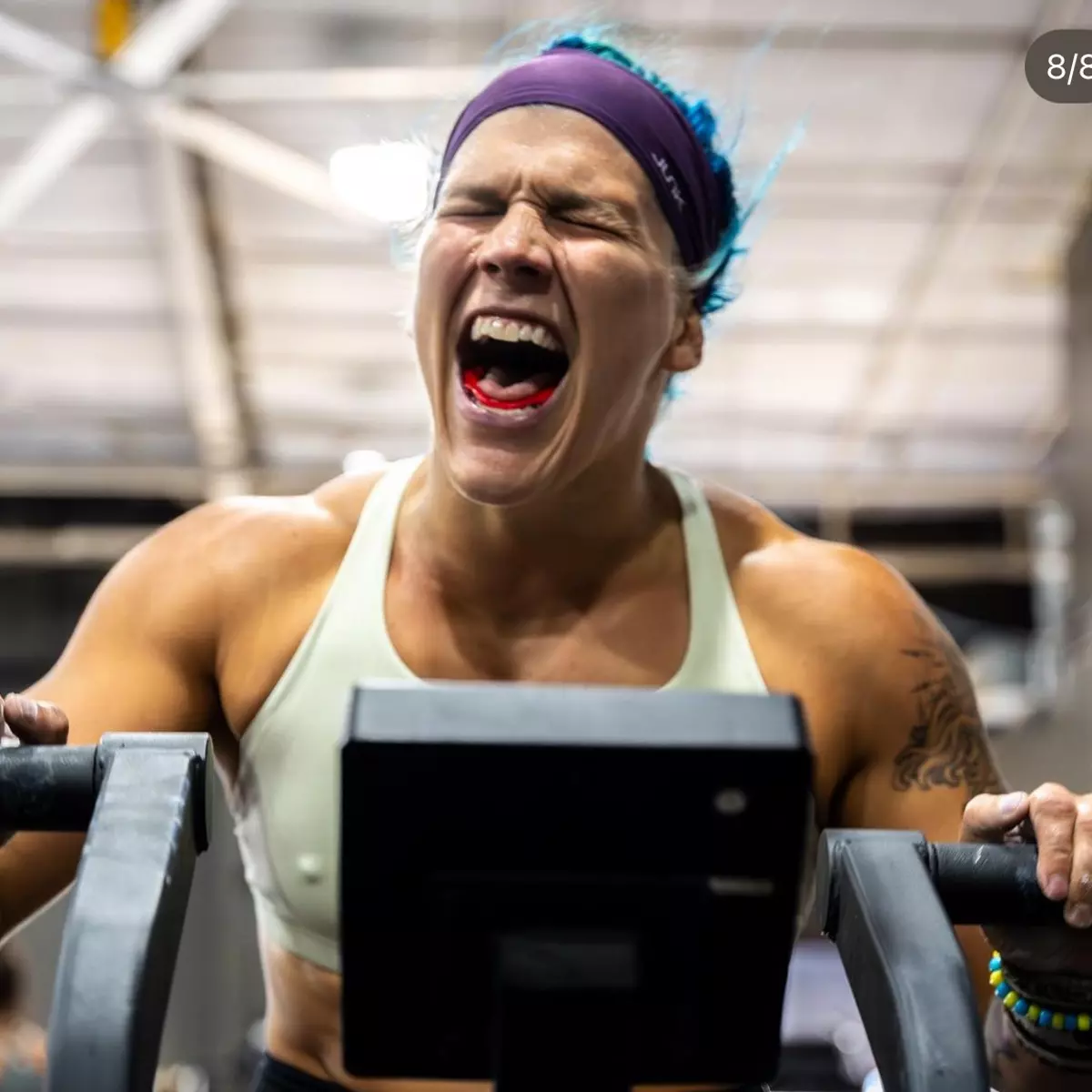 A woman with blue hair yells in exertion while using an exercise machine in a gym.