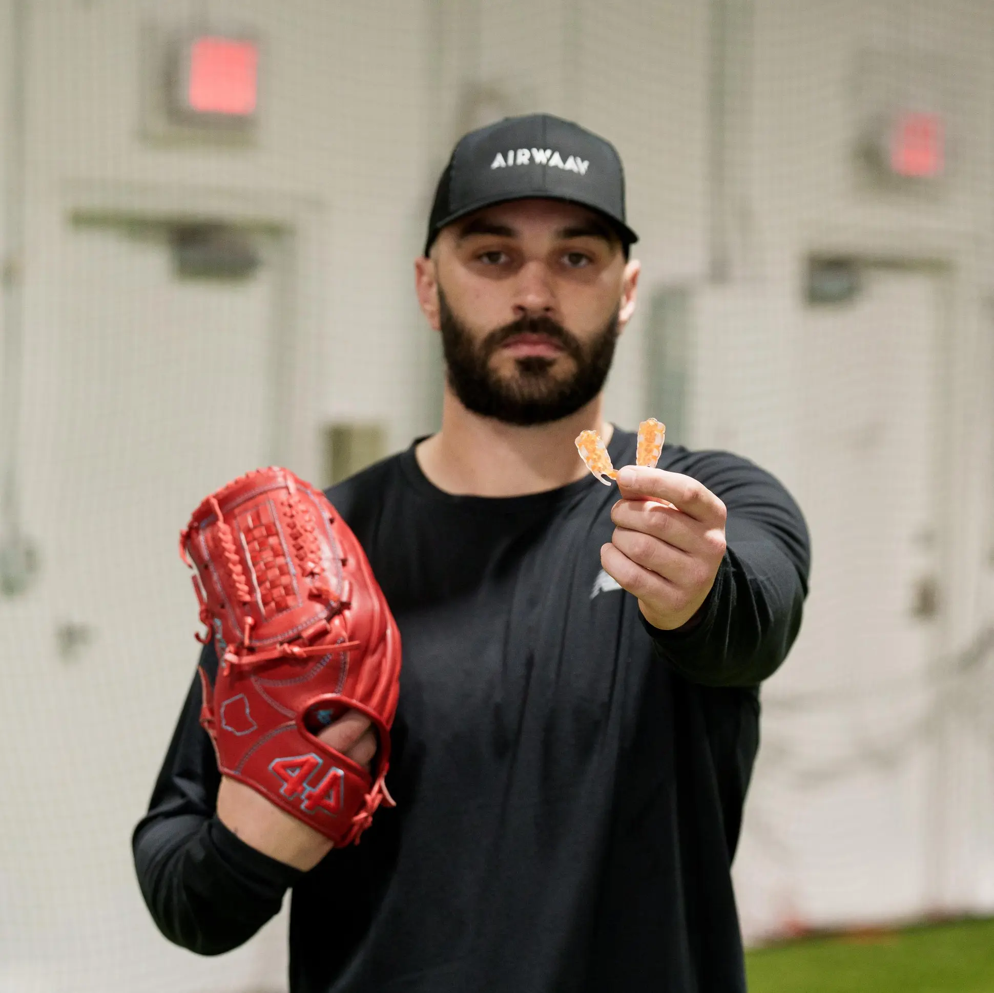 A baseball player in a black shirt and hat holds a red glove and a mouthguard.