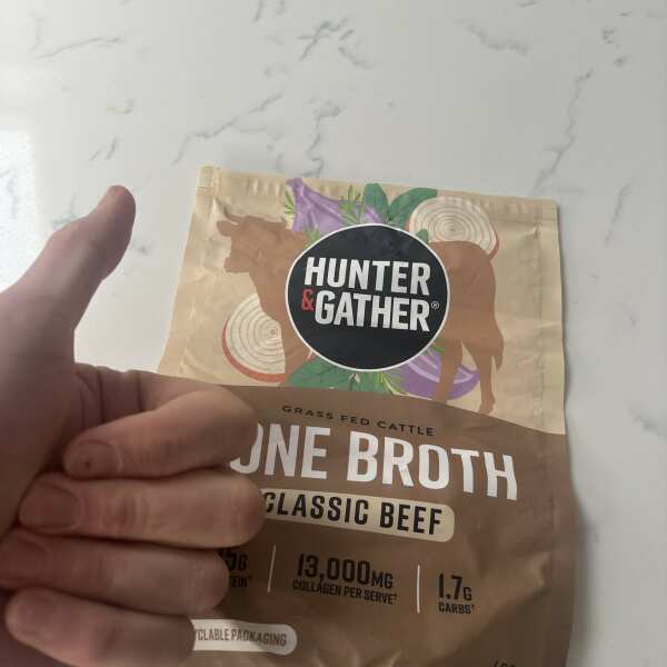 A person's hand holding a packet of Hunter & Gather Classic Beef Bone Broth.