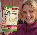 A smiling woman holds a bag of Grubterra Hen Harmony feed in front of a barn with chickens.