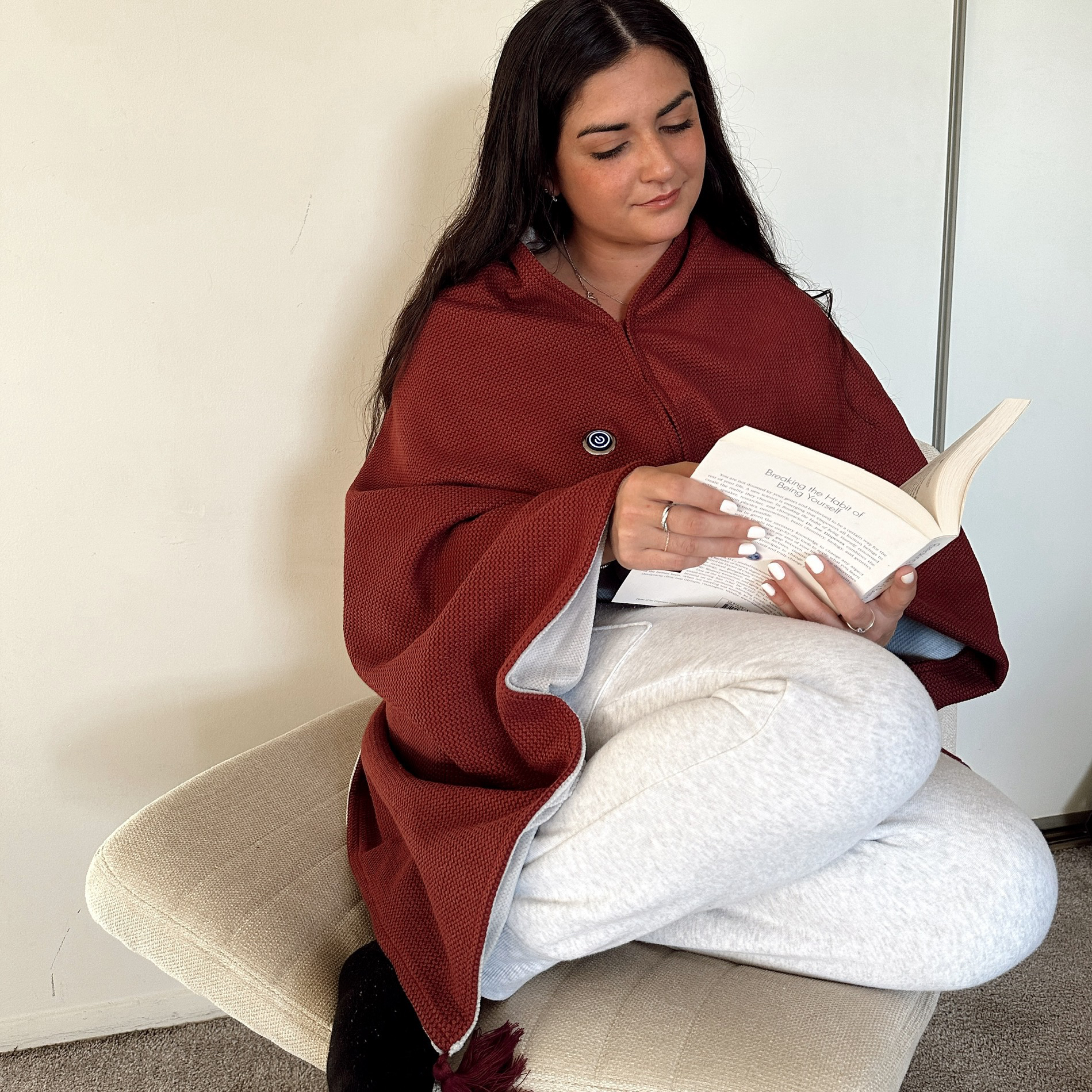 Person reading a book, sitting on a bench, wearing a red blanket and light-colored pants.