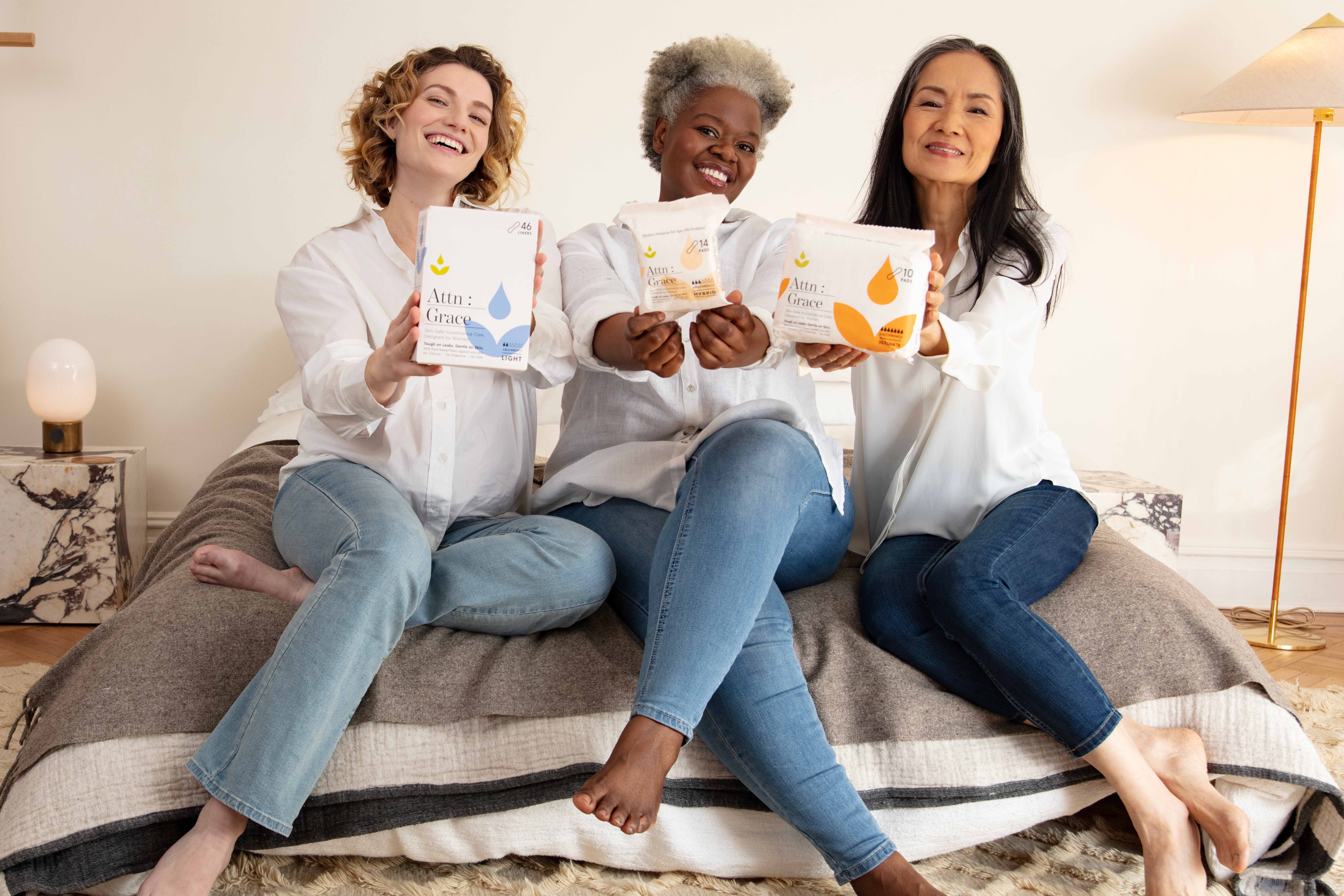 Three diverse women sit smiling on a bed, holding up packages of Attn: Grace absorbent products.