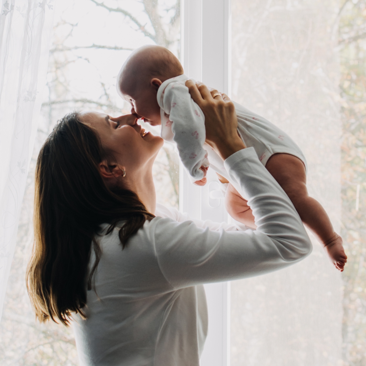 A woman lifts a smiling baby near a window.