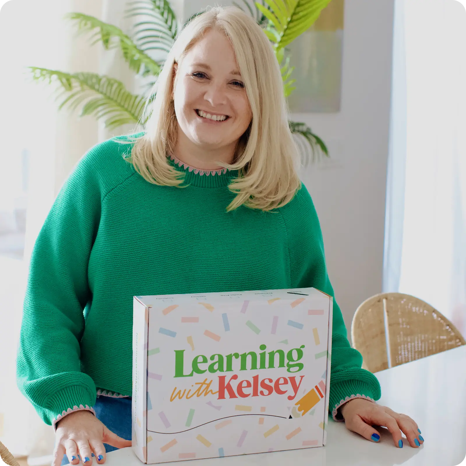 A smiling blonde woman in a green sweater with a box that says 'Learning with Kelsey' on a table.