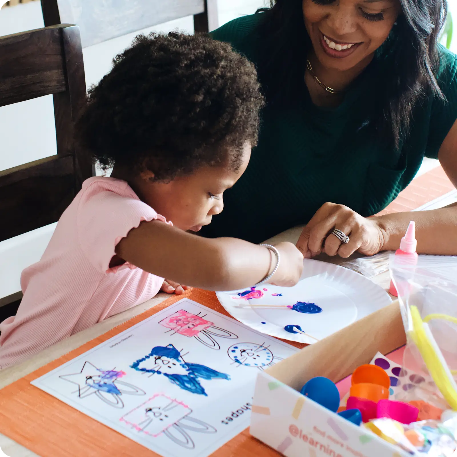 A woman smiles while helping a young child with a painting craft project at a table.