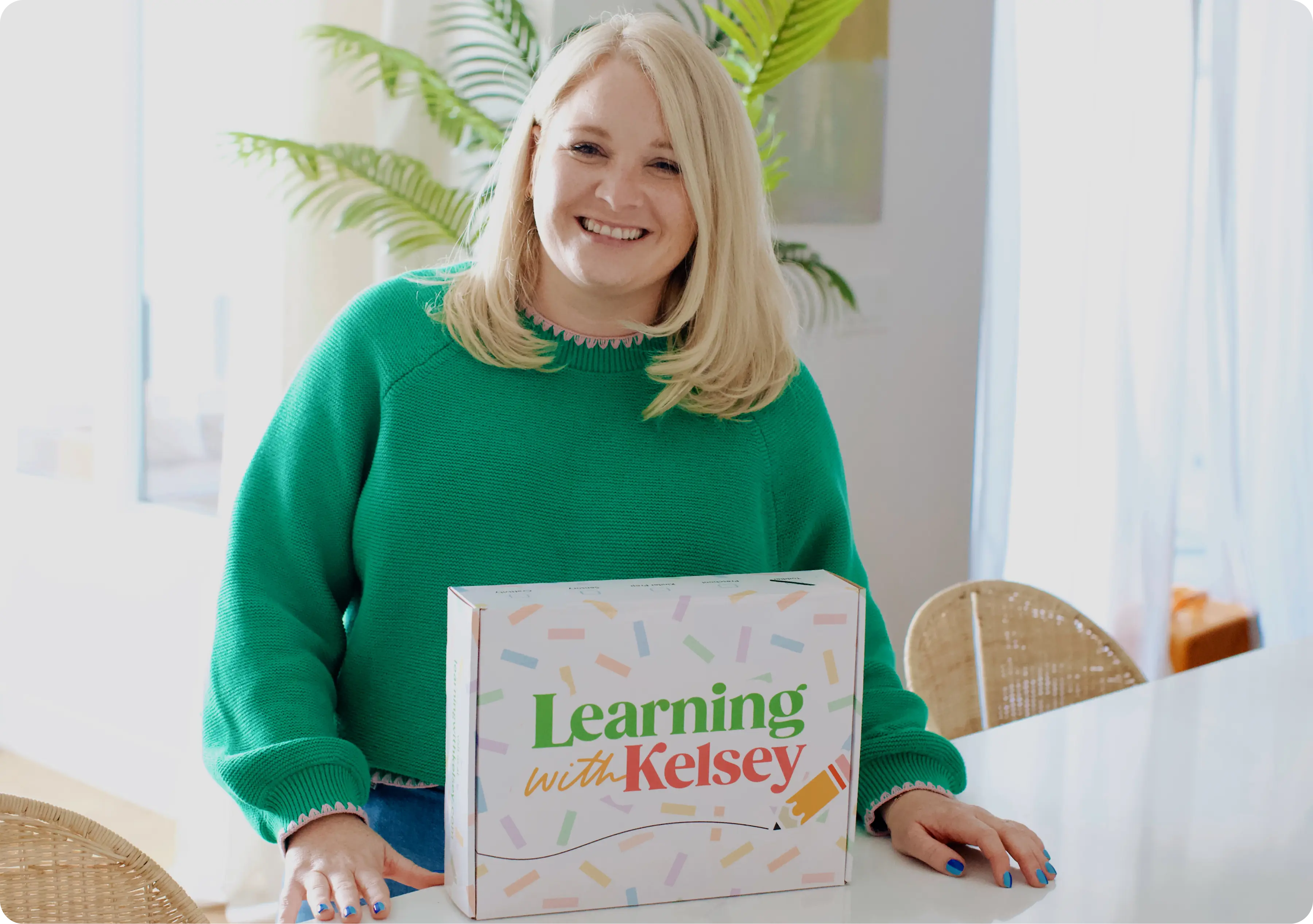 A smiling woman in a green sweater sits at a table with a 'Learning with Kelsey' box.