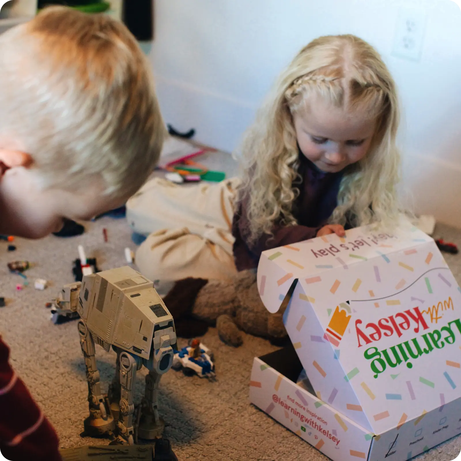Two young children play on a carpet with toys and an open 'Learning with Kelsey' box.