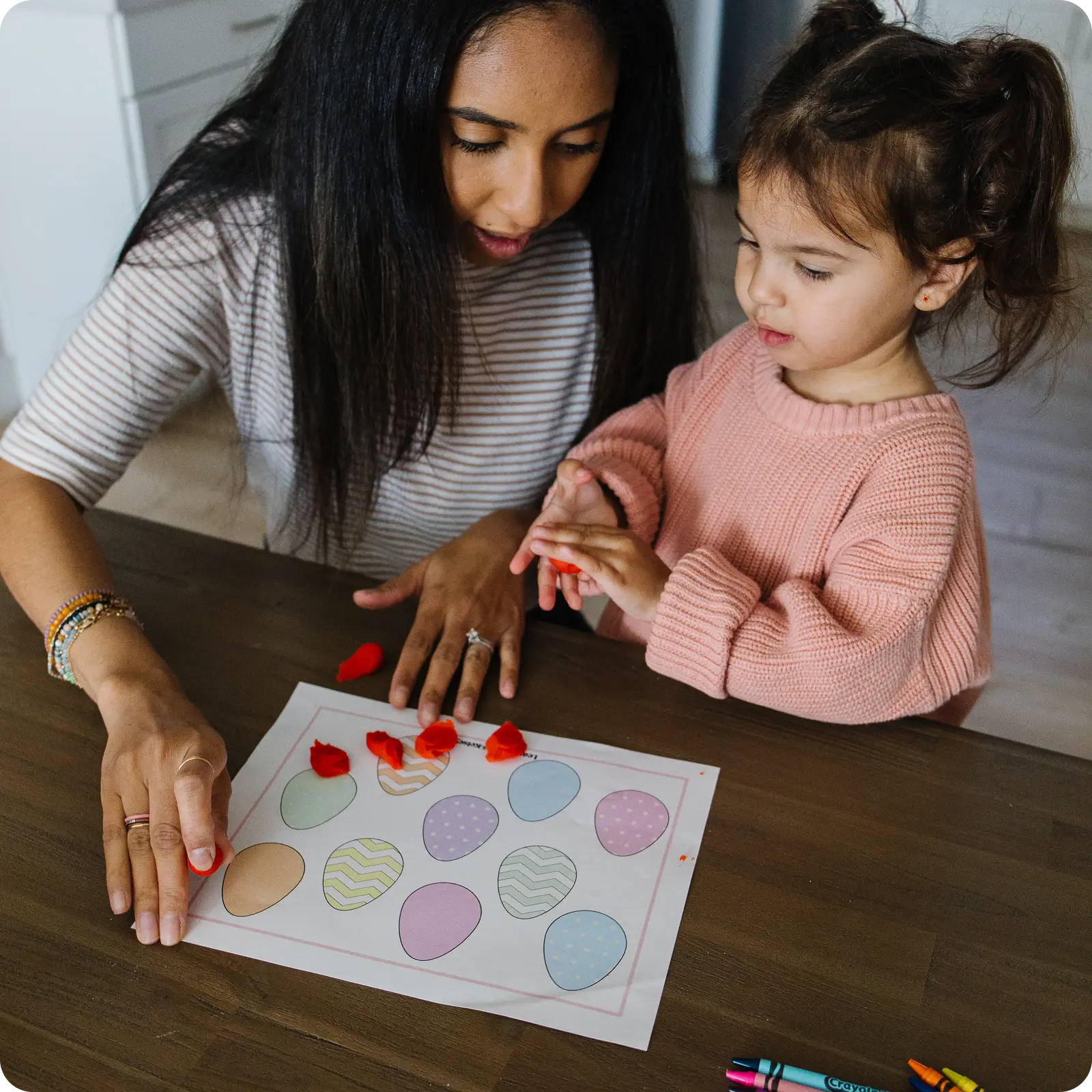 A woman and a young child sit at a table doing an arts and crafts activity with paper eggs.