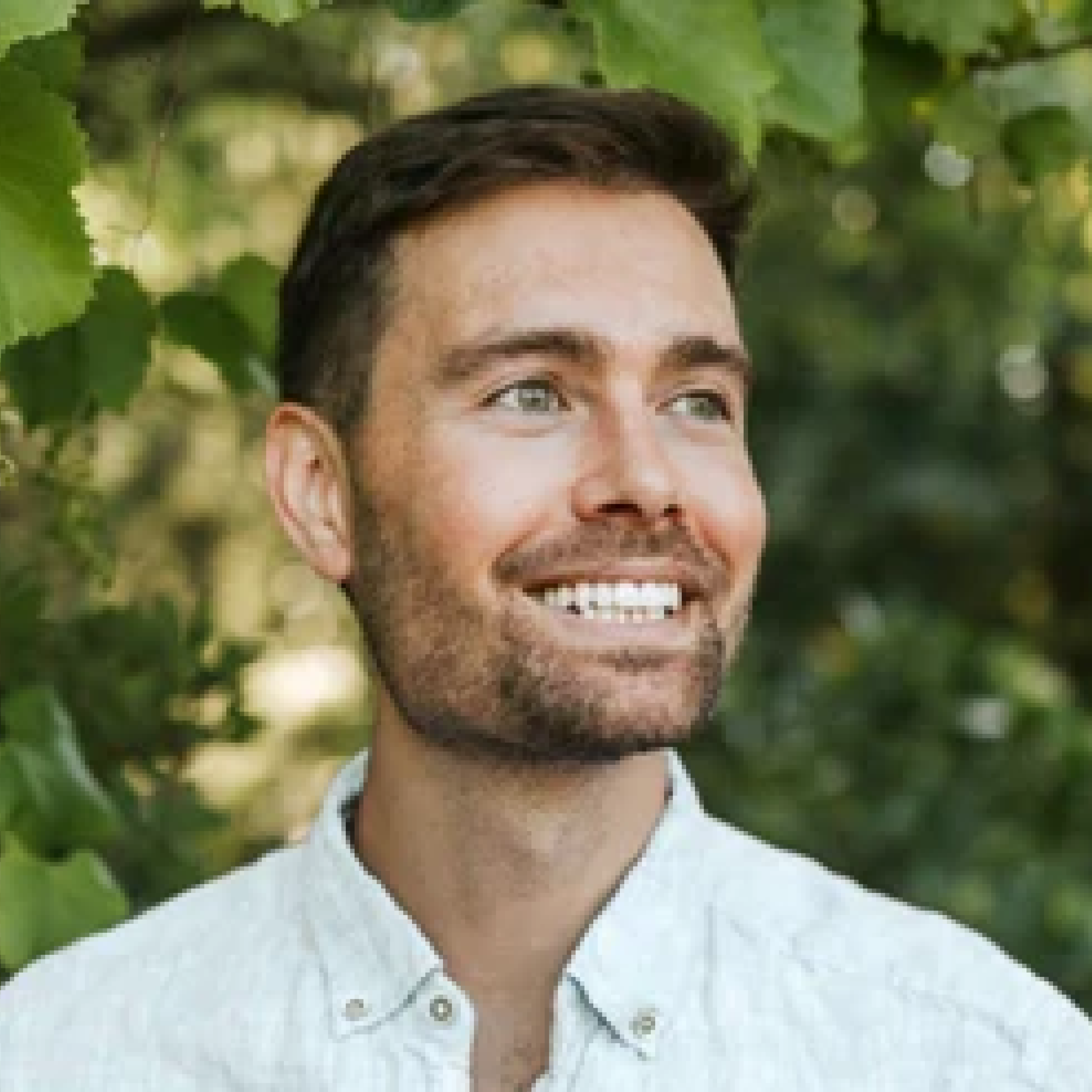 A headshot of a smiling man with a beard, looking to the side with a background of green leaves.