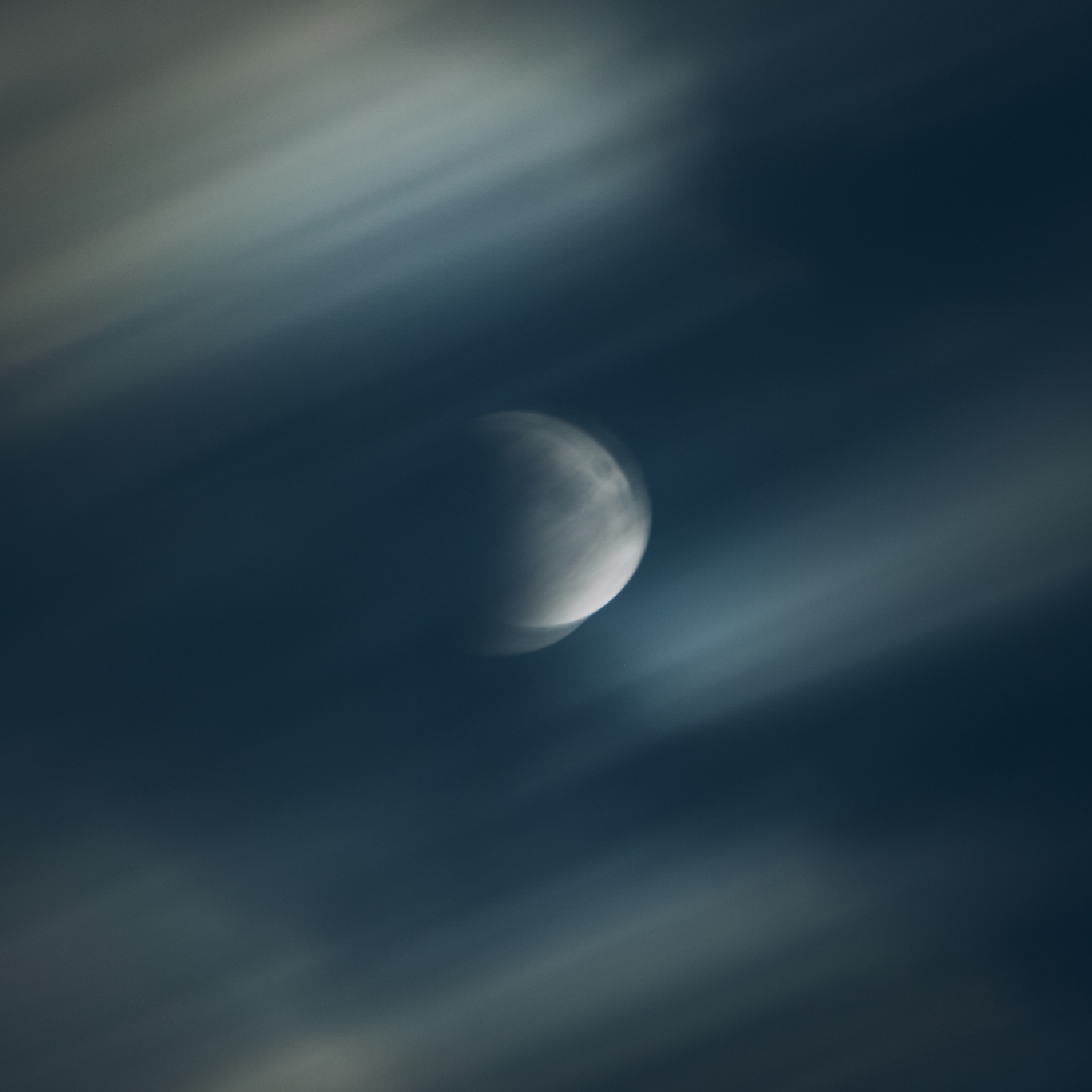 A long exposure photograph of a gibbous moon partially obscured by streaky, motion-blurred clouds.
