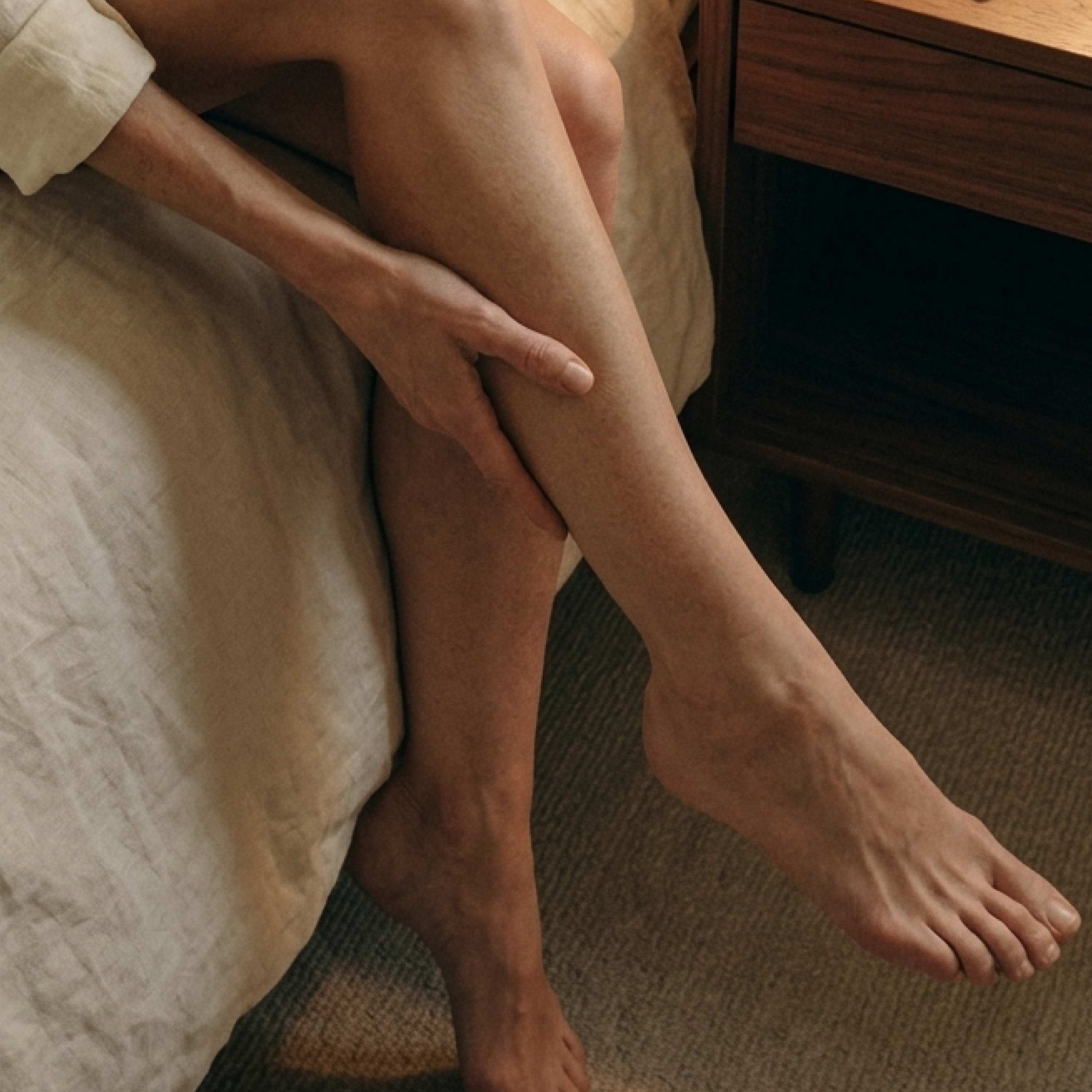 A person sits on the edge of a bed, with their hand resting on their lower leg.