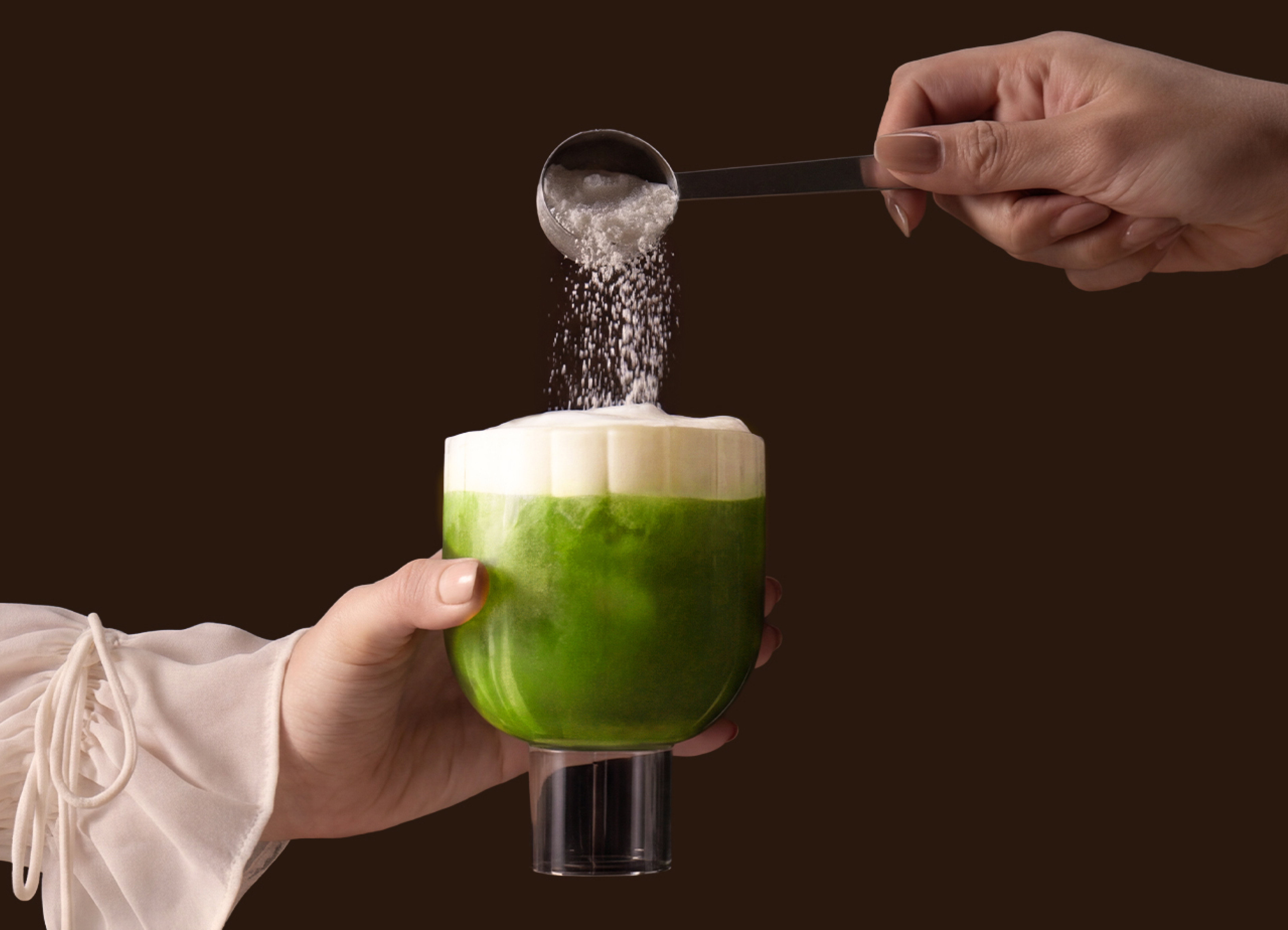 A hand pours powder from a measuring spoon onto a foamy green drink held in a glass.