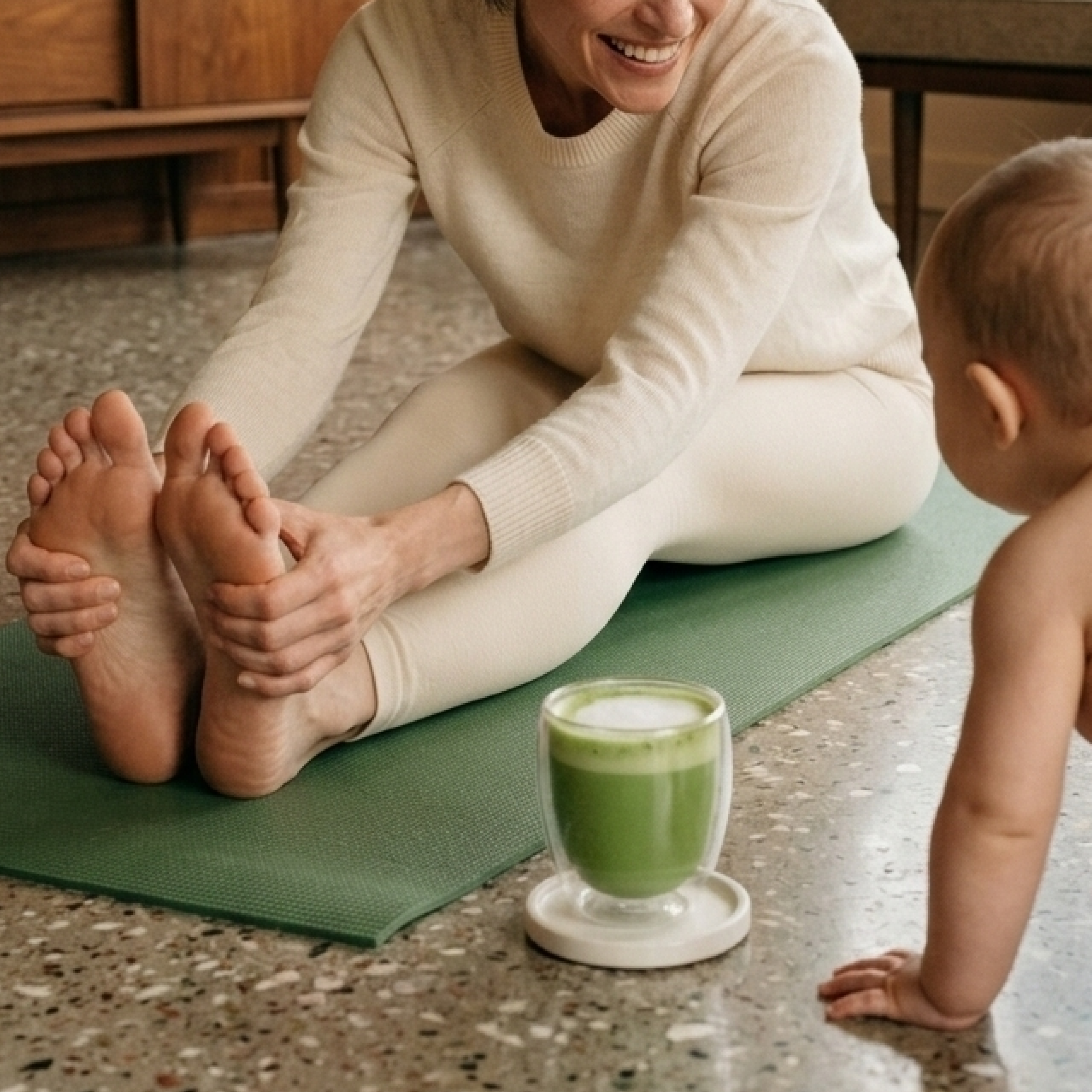 A woman stretches on a yoga mat, holding her feet, as a baby watches her from the floor.