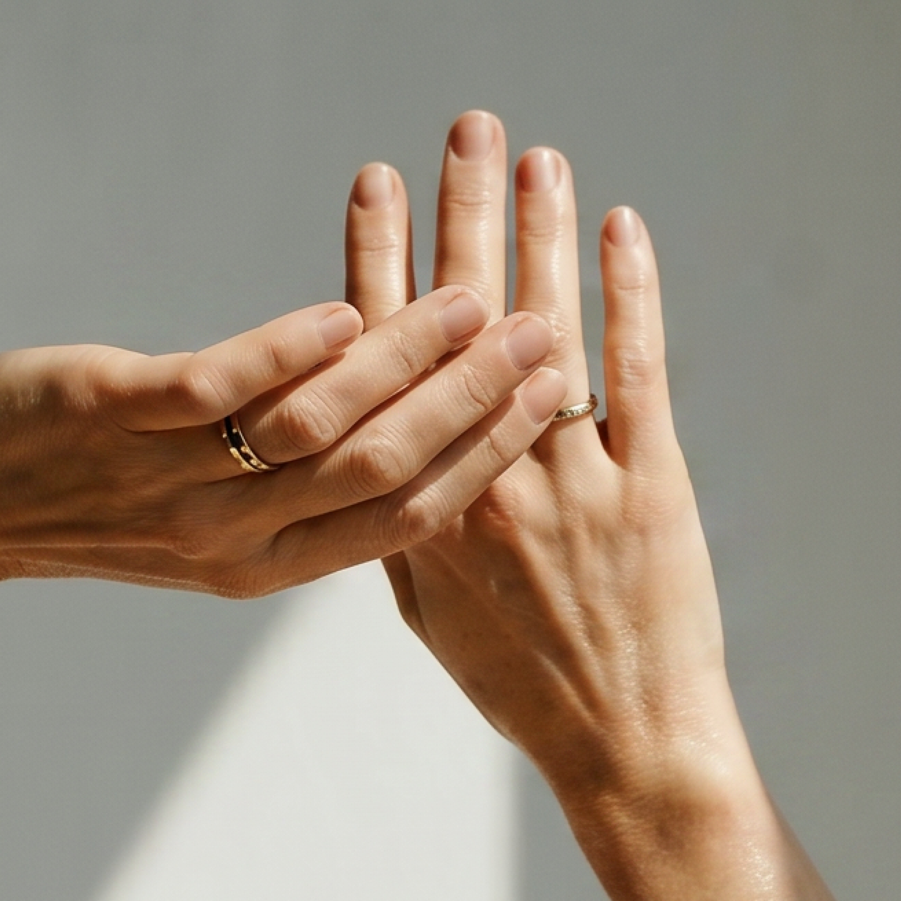 A close-up of two hands gently holding each other, both wearing rings, against a plain background.