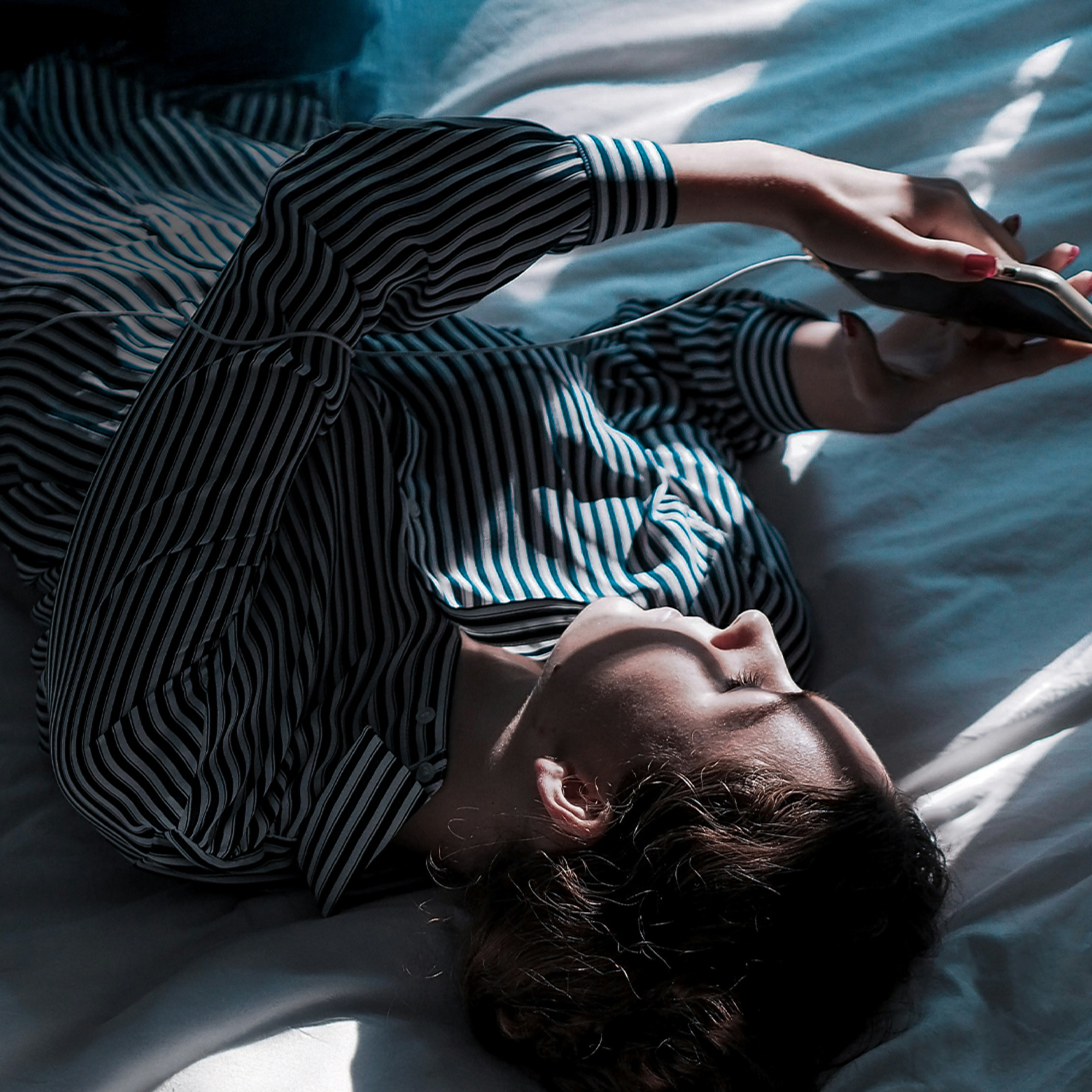 A person in a striped shirt lies down, holding a smartphone with wired earphones.