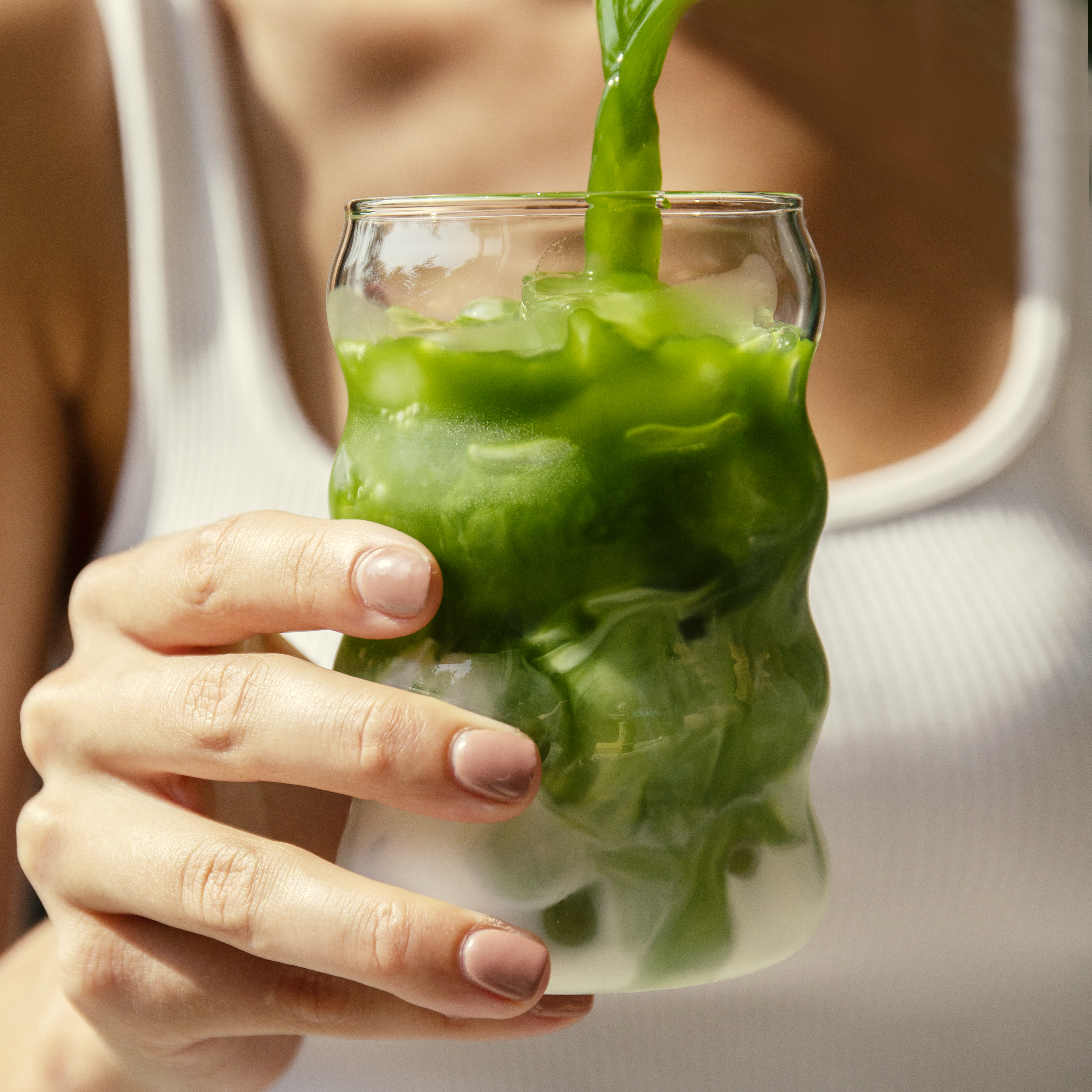 A person pours green matcha into a wavy glass of iced milk.