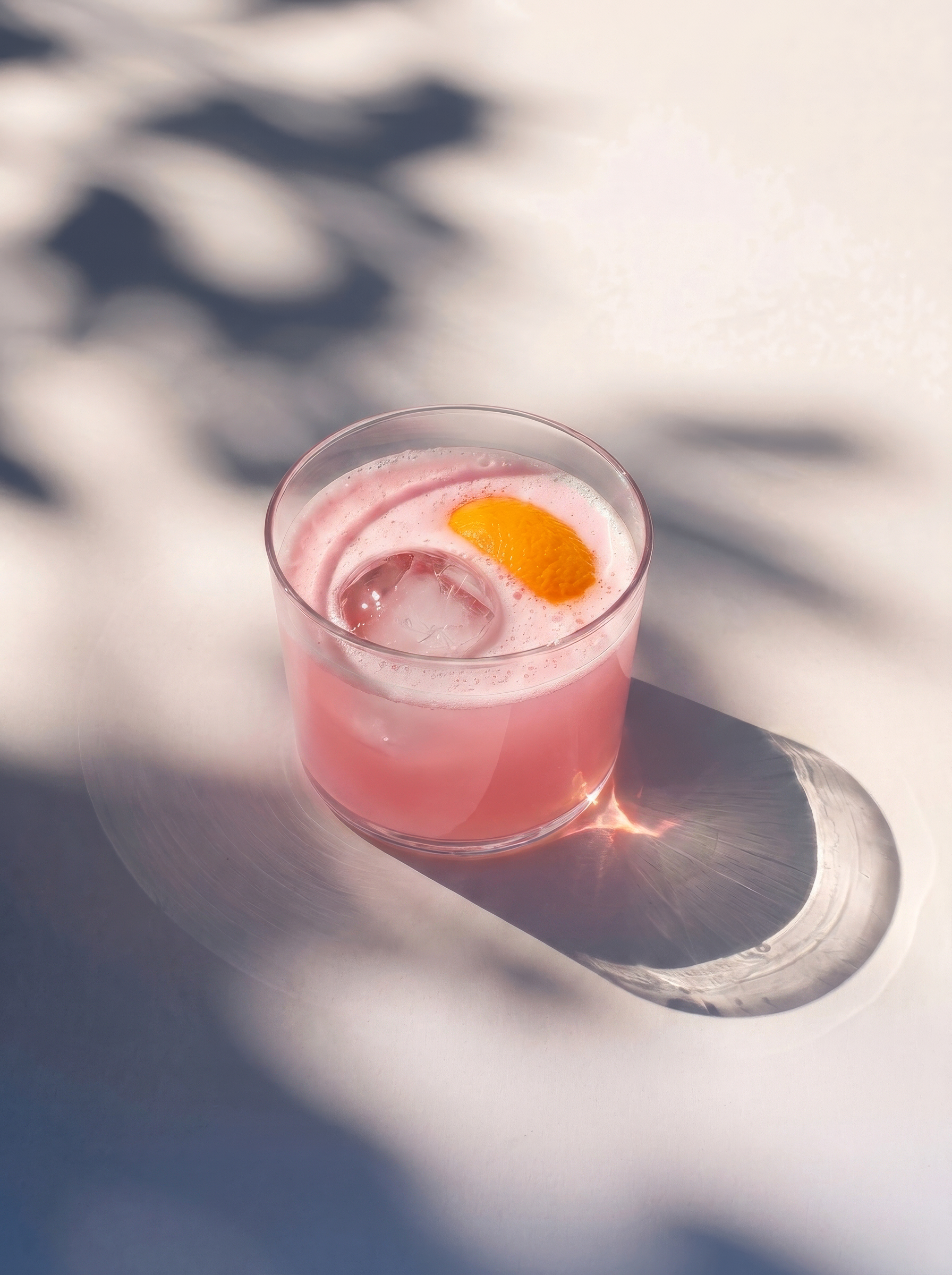 A pink cocktail in a short glass with an orange peel garnish, sitting in dappled sunlight.