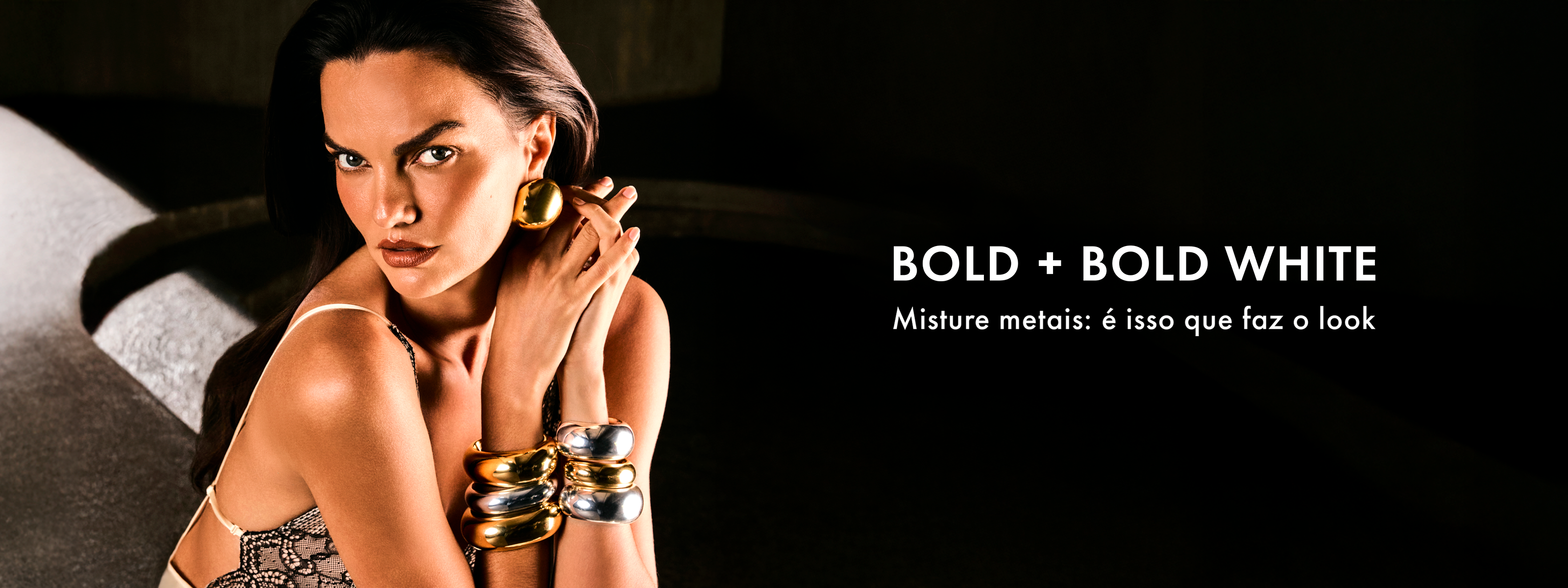 A woman looks intensely at the camera, wearing bold, mixed-metal gold and silver earrings and bracelets.
