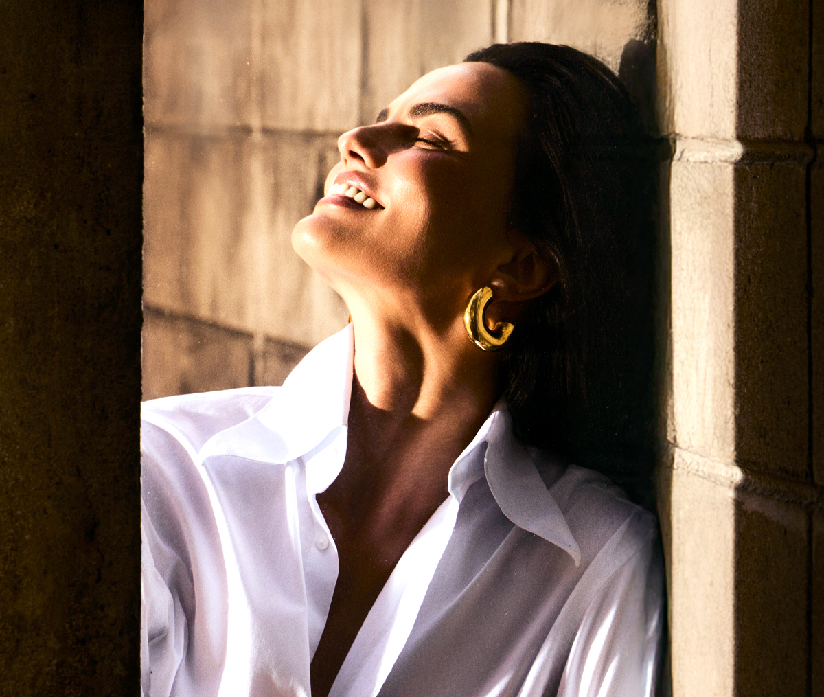 A woman in a white shirt leans against a wall, smiling with her face in the sun.