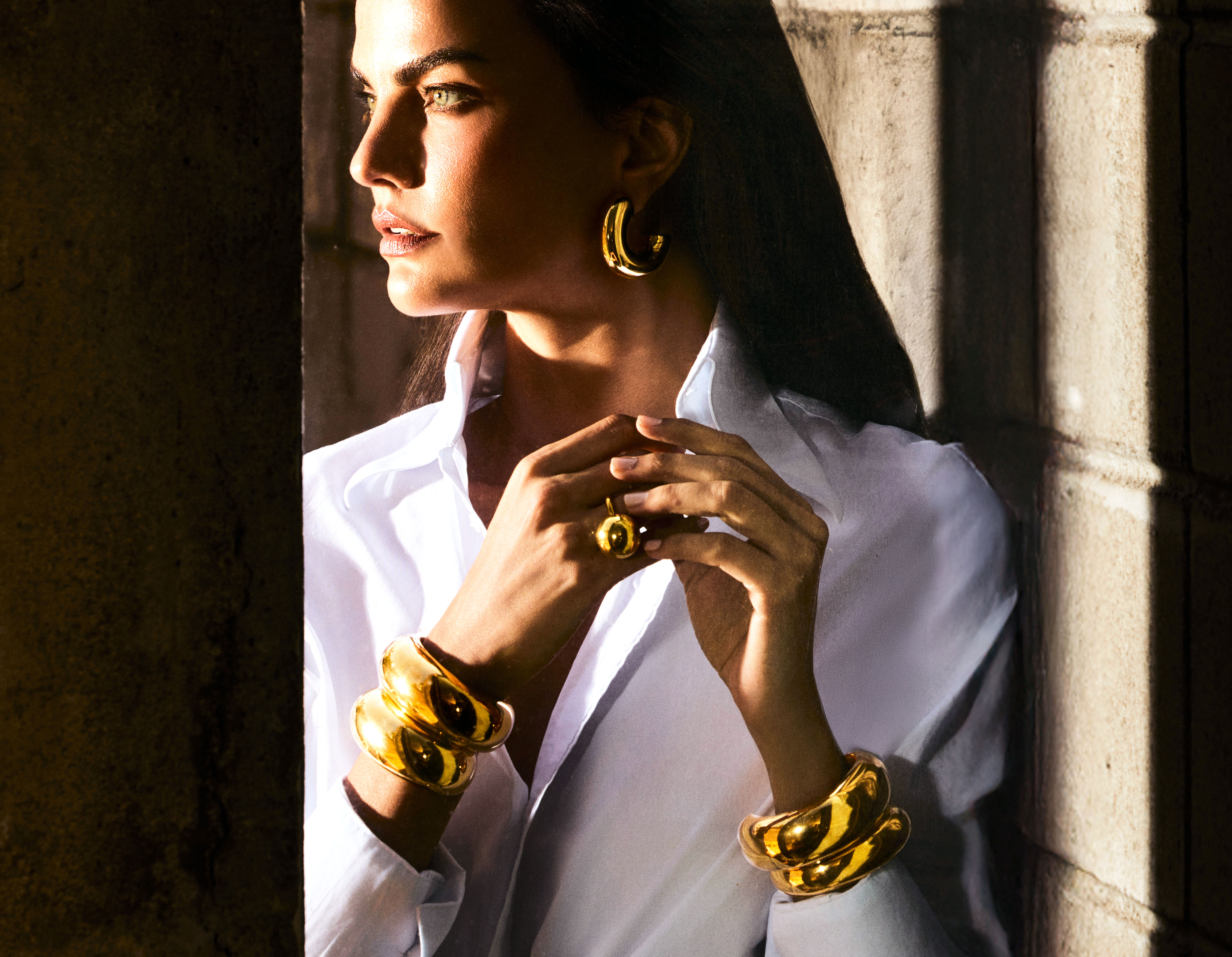 A woman in a white shirt and bold gold jewelry stands in dramatic light, seen in profile.