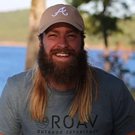 Man with long hair and beard wearing a cap, smiling outdoors.