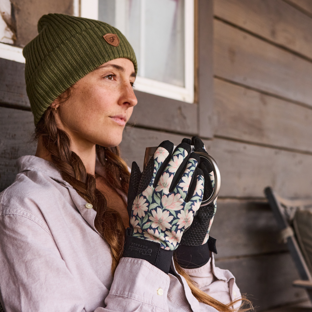 A woman in a green beanie and floral gloves holds a metal mug while looking off to the side.