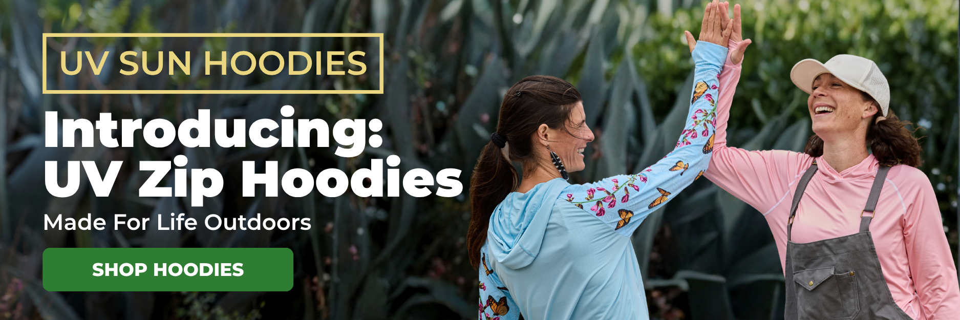 Two smiling women in sun hoodies give each other a high-five outdoors in front of leafy plants.