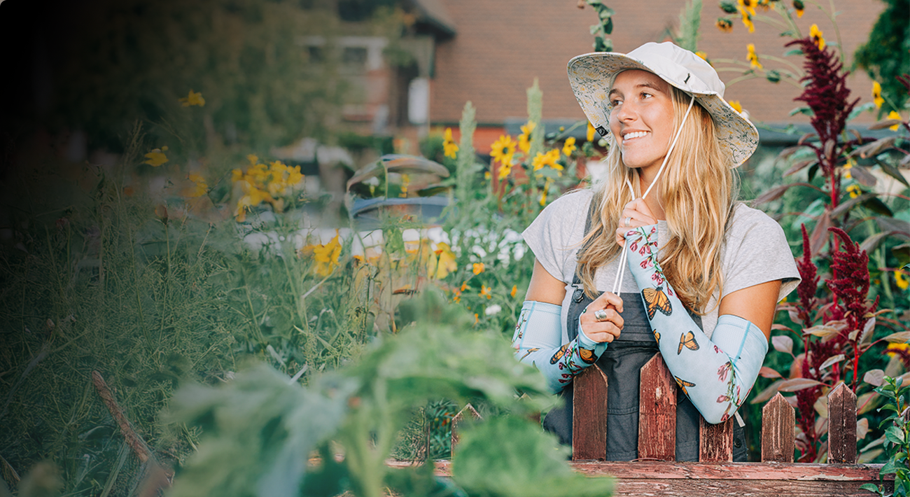Woman in a garden wearing a sun hat and floral sleeves, surrounded by flowers.