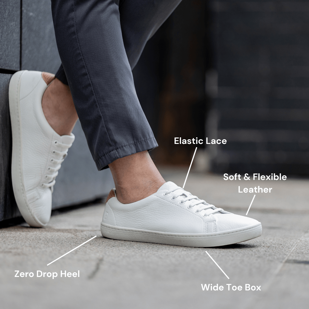 Person wearing white sneakers with labeled features: elastic lace, soft leather, wide toe box, zero drop heel.