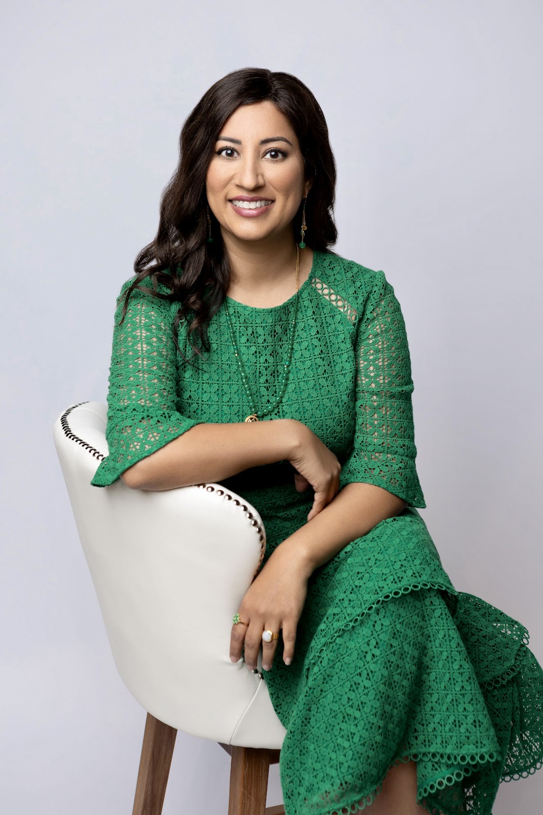 A woman with dark hair in a green lace dress sits in a white chair and smiles.