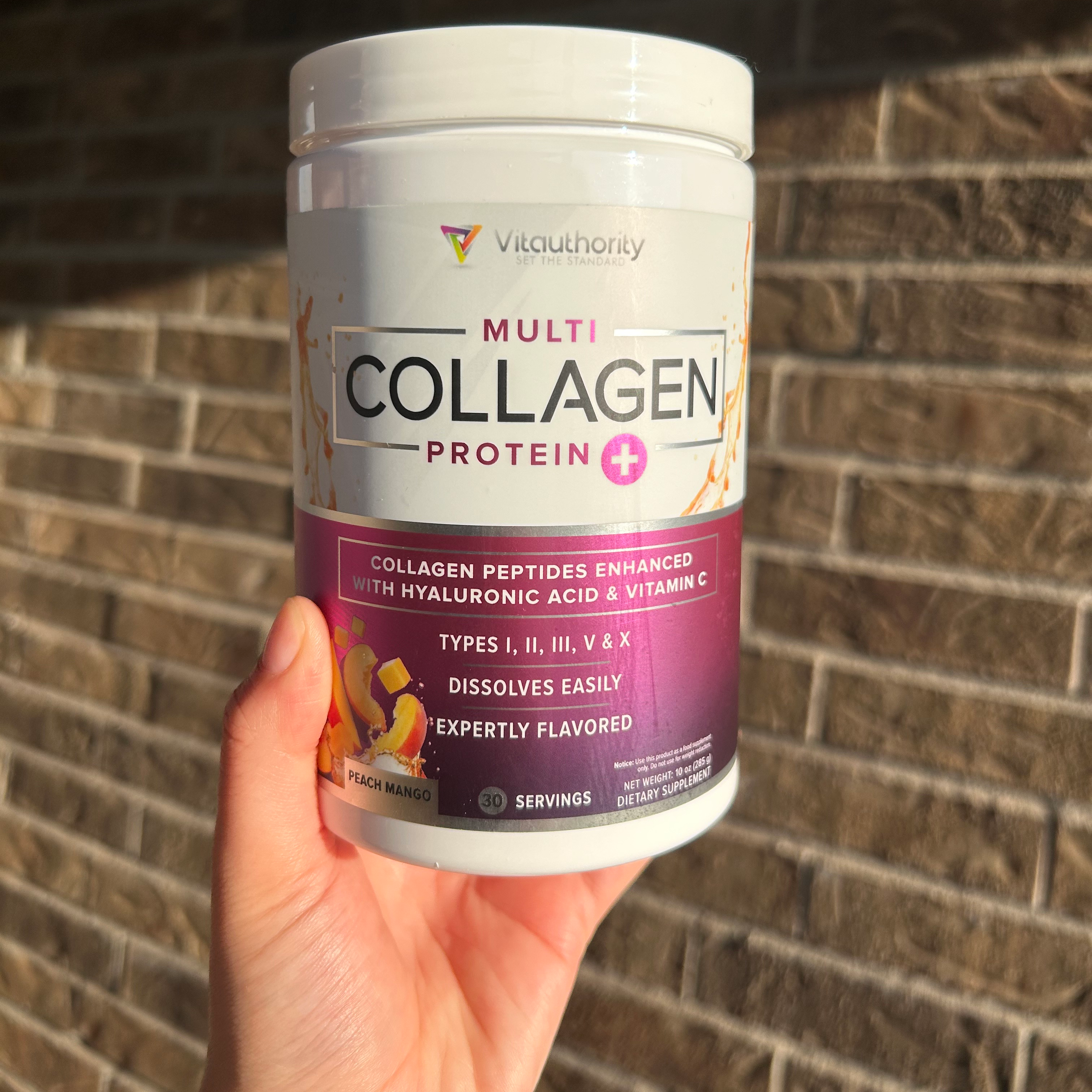 A hand holding a white container of Vitauthority Multi Collagen Protein+ against a brick wall.