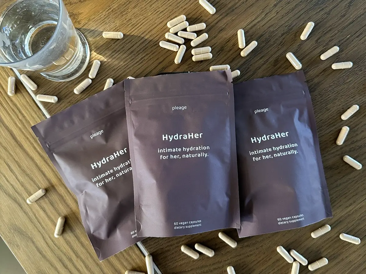 Three packets of HydraHer supplements surrounded by capsules on a wooden table.