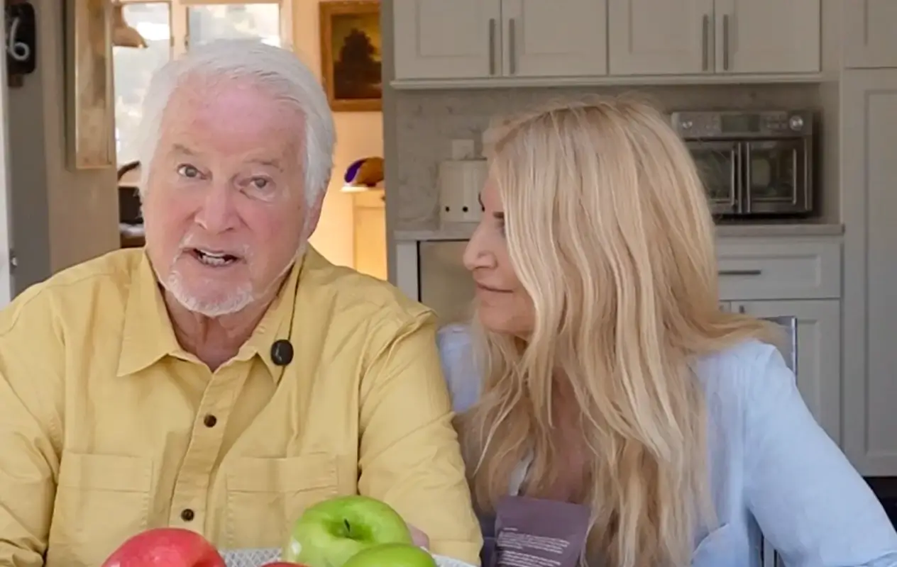 An older man and woman in a kitchen with apples on the table.